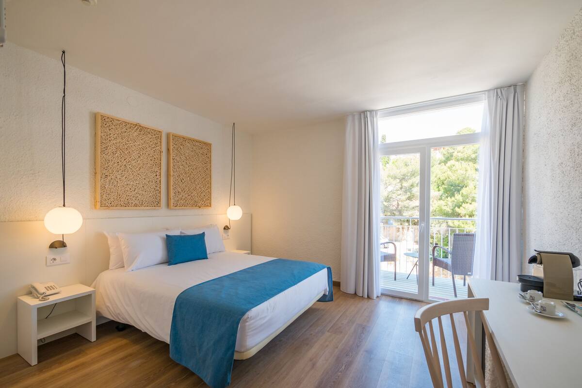 A hotel room with a bed, bedside table, and a desk with a chair. There is a balcony with a table and chairs.