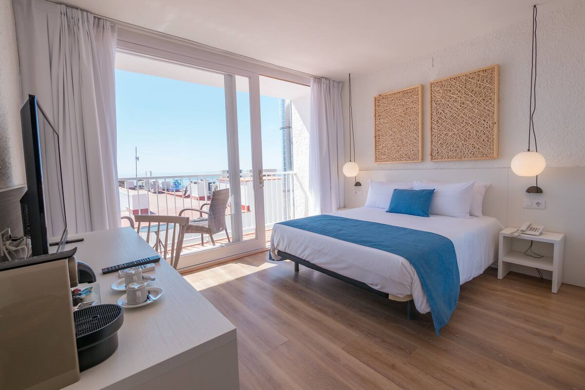 A hotel room with a bed, bedside table, and a desk with a computer and phone.
