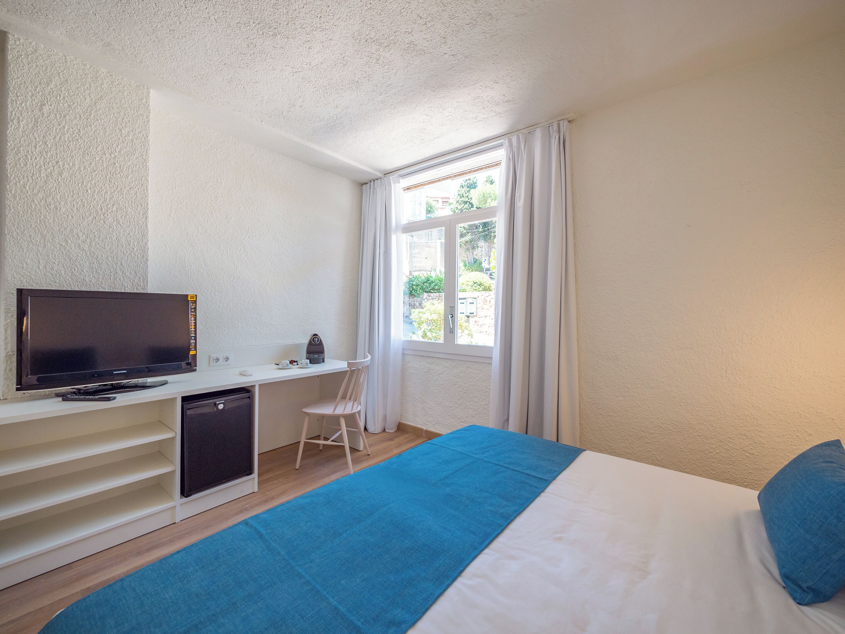 Un dormitorio con una cama, un escritorio con televisor, una silla y un ventanal con cortinas.