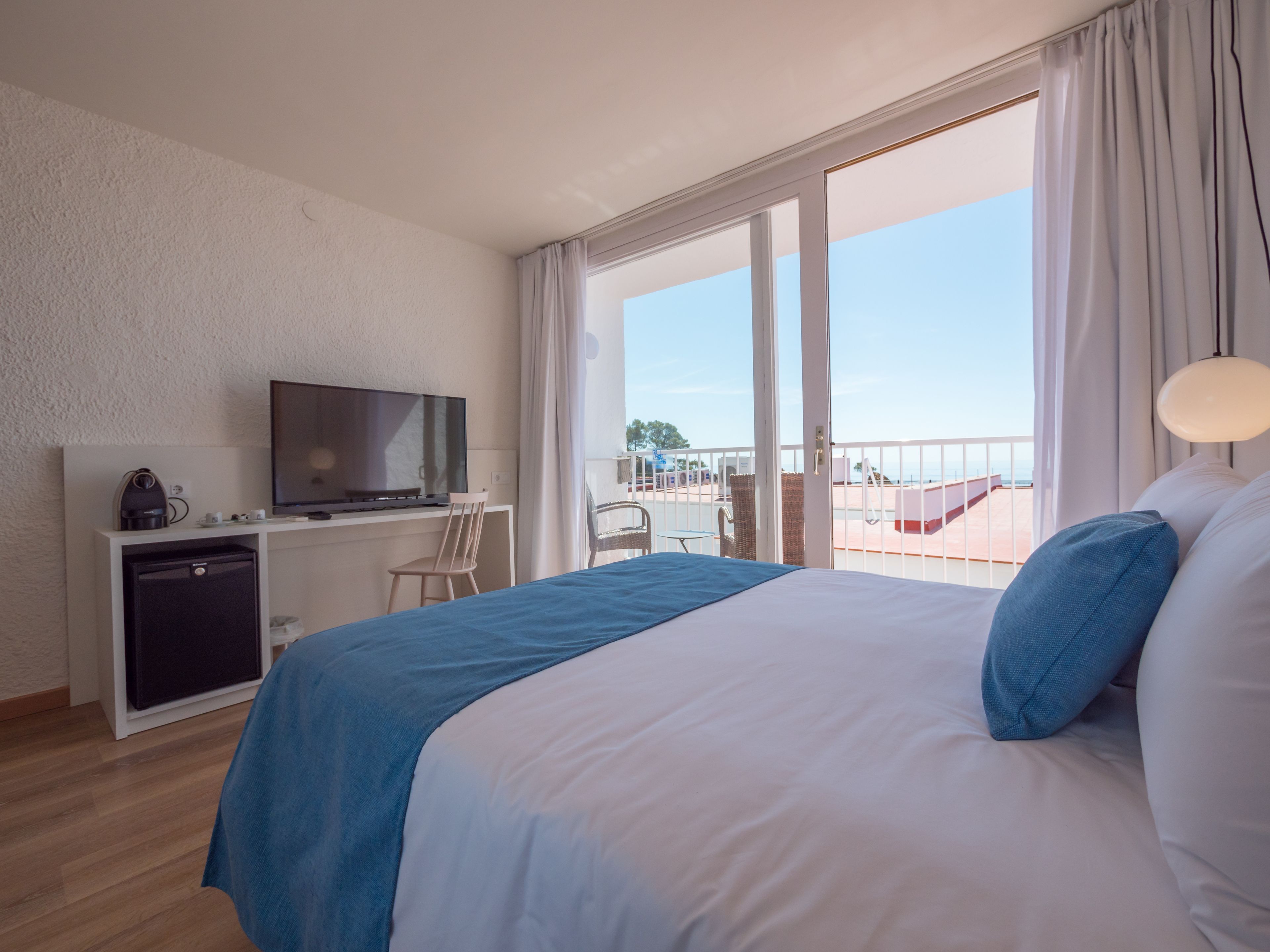 A hotel room with a bed, a desk with a television, a small fridge, and a balcony with a view of the sea.