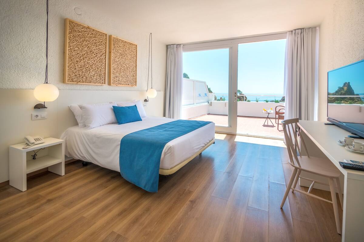 A hotel room with a bed, a desk, a TV, and a balcony with a view of the sea.