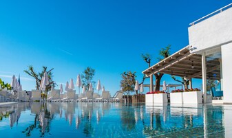 Uma área de piscina com espreguiçadeiras e guarda-sóis, um espaço de estar coberto e um céu azul límpido.
