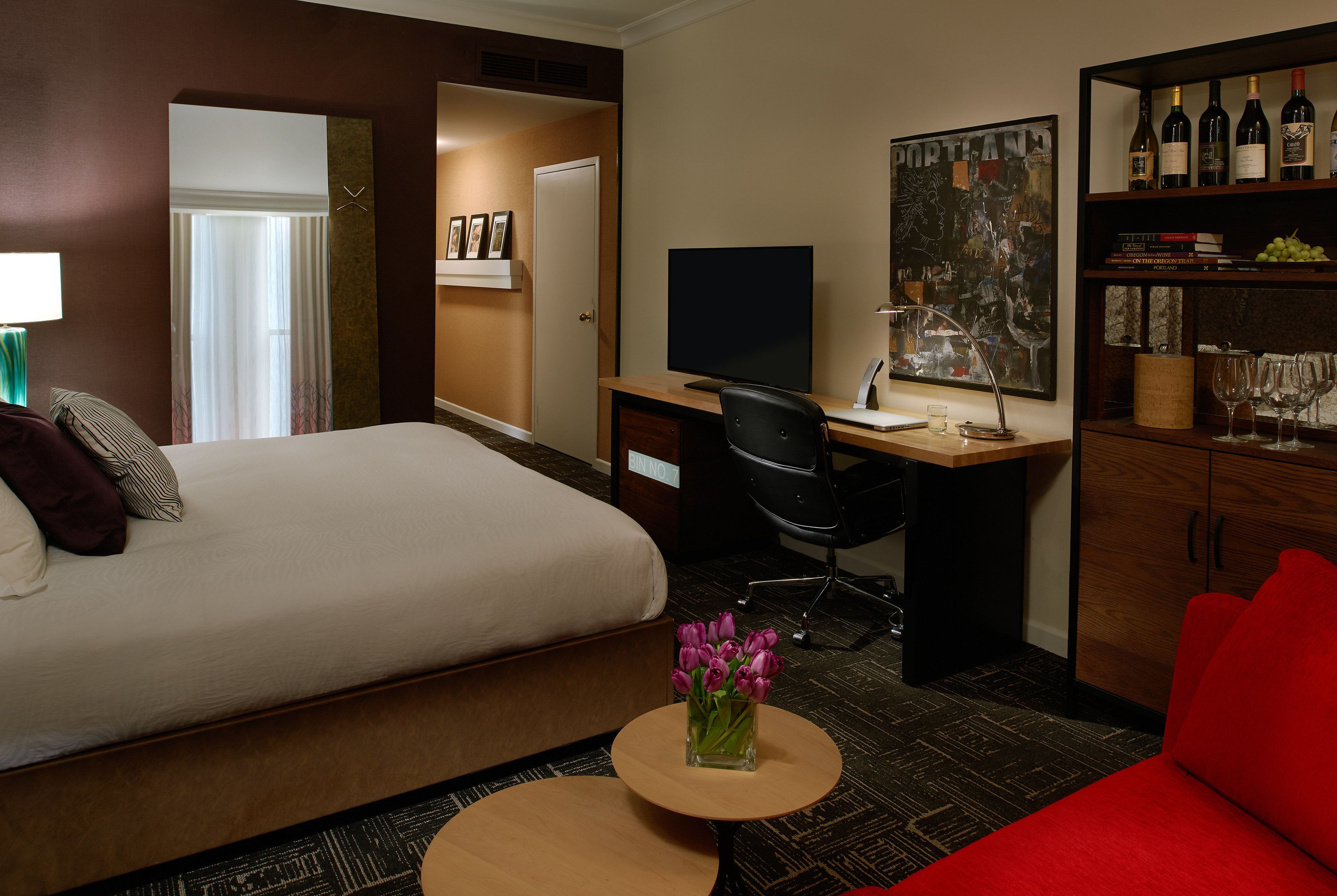 Habitación de hotel con cama, escritorio con computadora, silla, mesita con un florero, sofá rojo y estantería con botellas de vino.