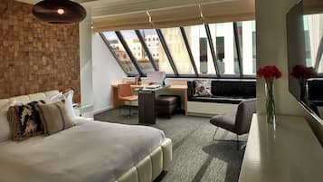 Quarto de hotel moderno com uma cama grande, uma escrivaninha, uma cadeira e um sofá. Há uma janela ampla com vista para edifícios.