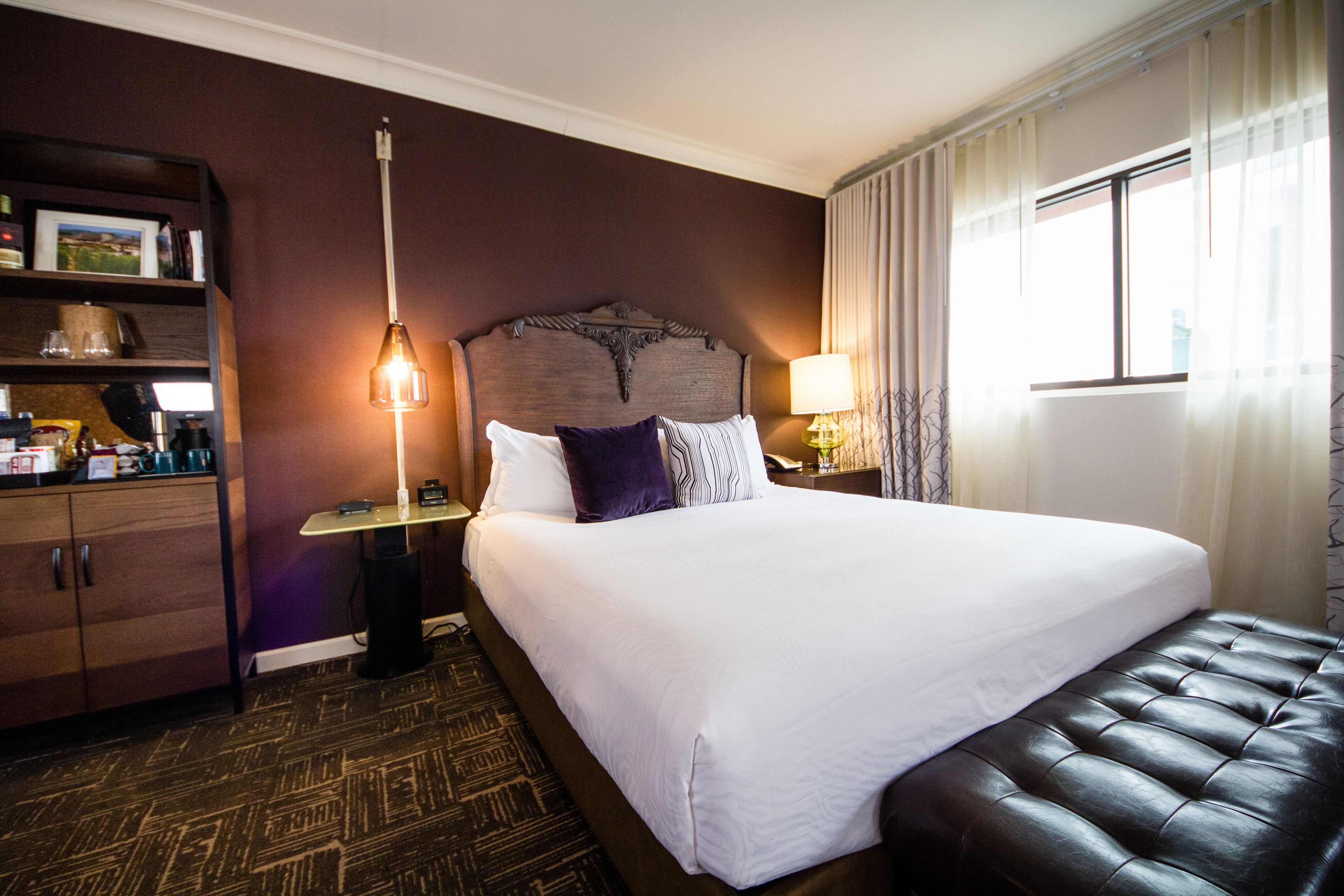 A hotel room with a large bed, a headboard, a nightstand with a lamp, a wooden cabinet, and a window with curtains.