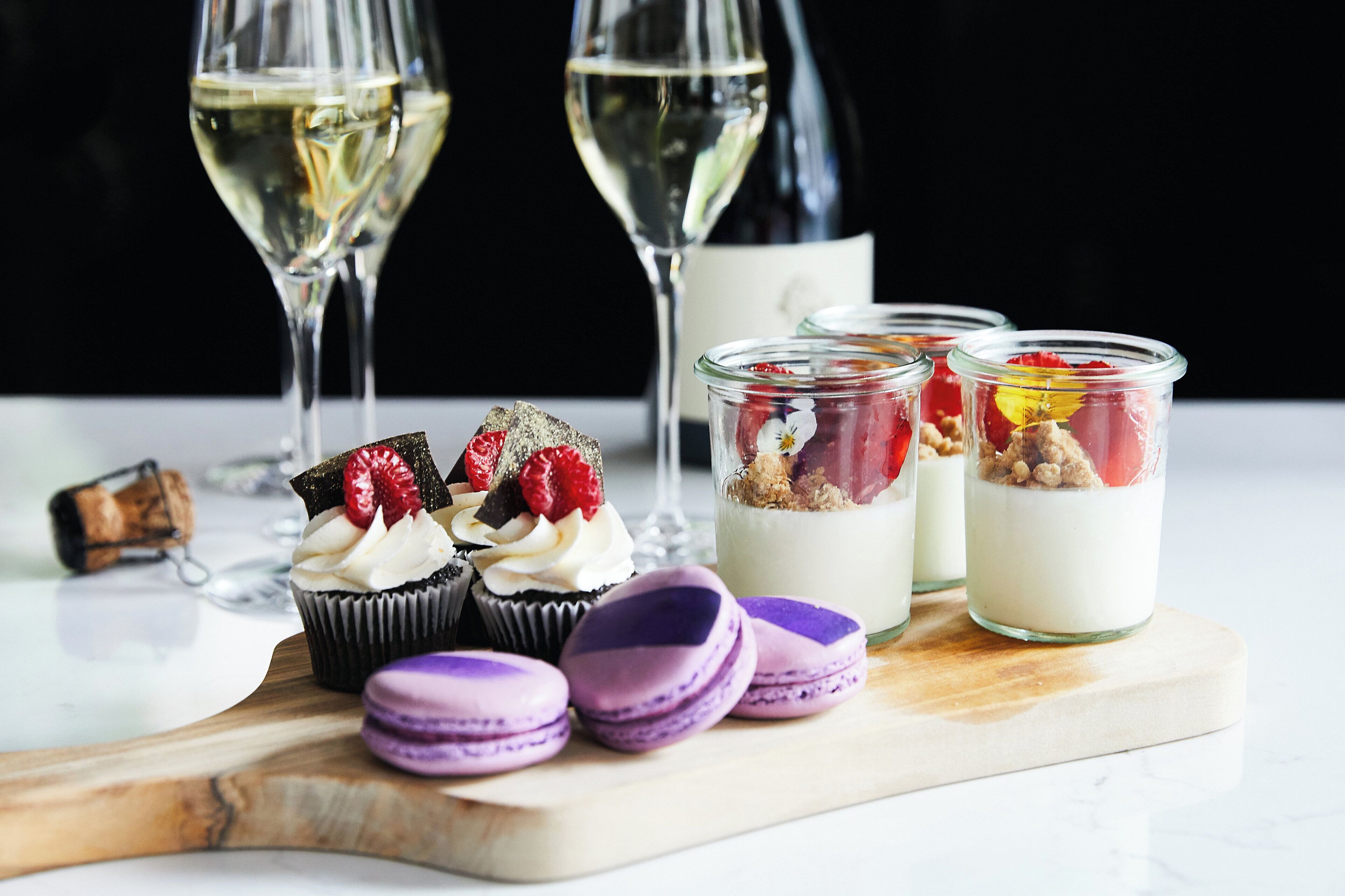 A dessert table with cupcakes, macarons, and layered desserts in glasses, with two wine glasses in the background.