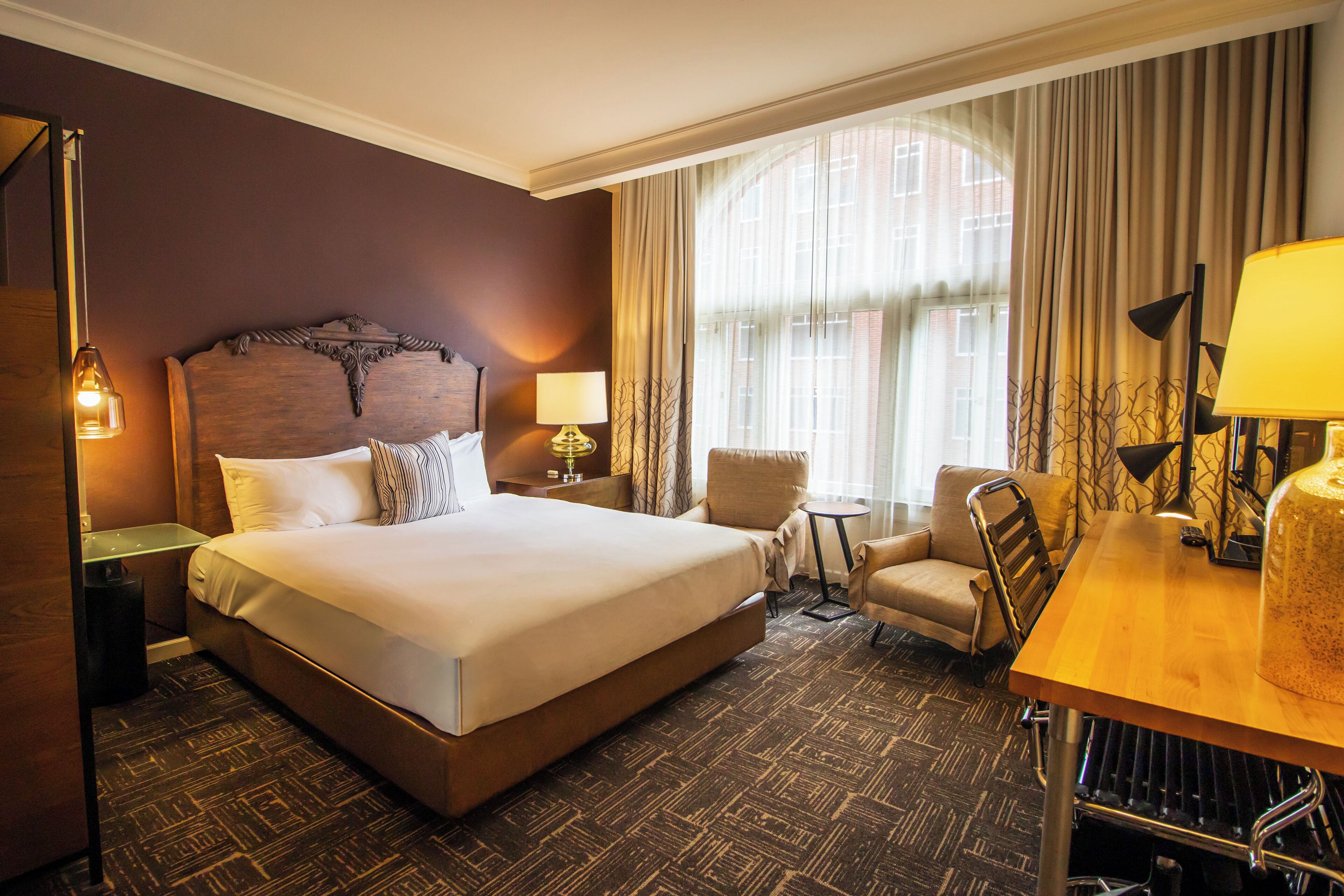 A hotel room with a large bed, a wooden headboard, a small table with a lamp, a chair, and a window with curtains.