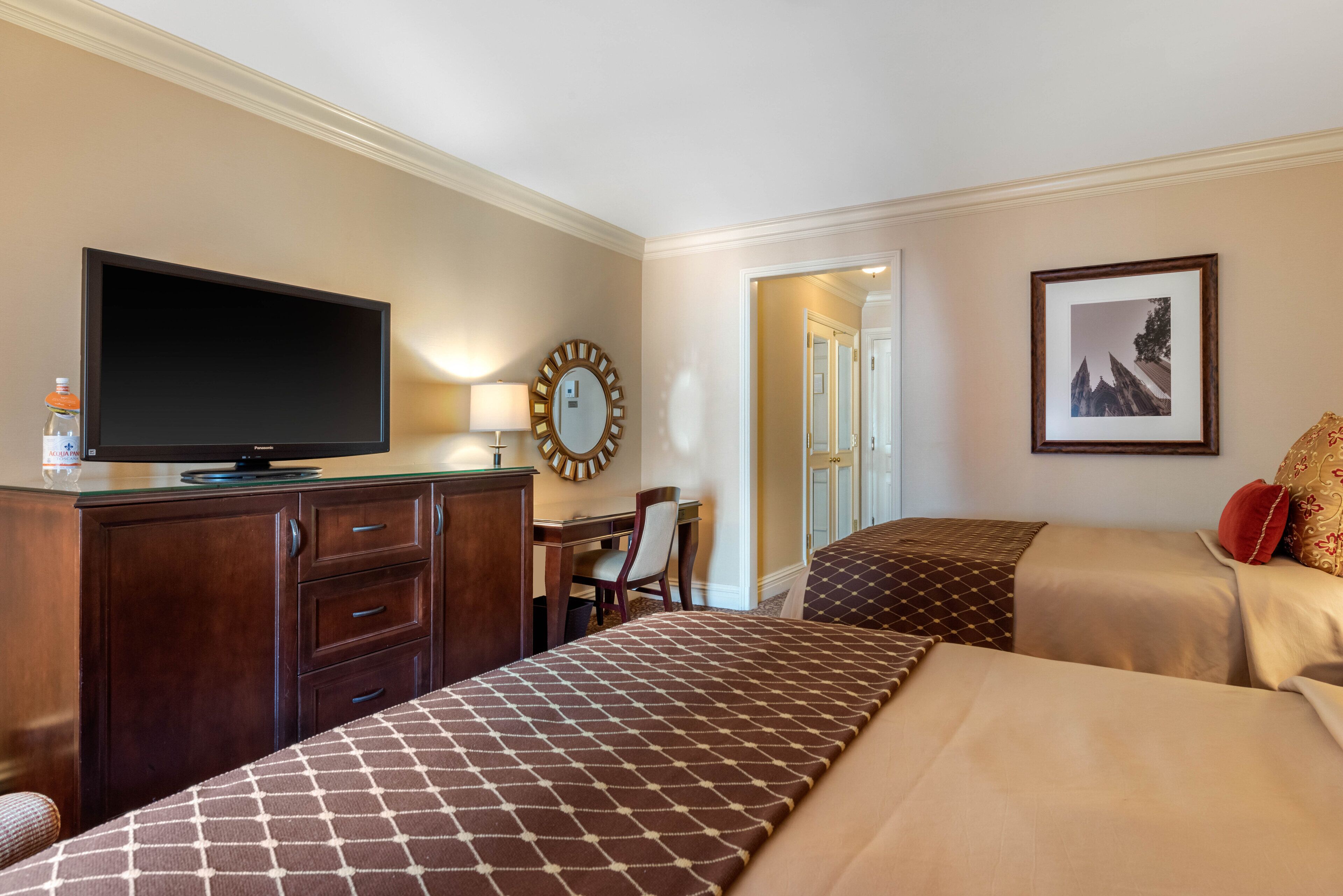 A hotel room with a TV, desk, chair, two beds, and a framed picture on the wall.