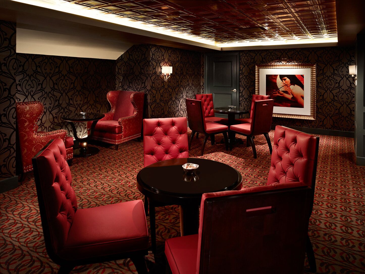 A room with red upholstered chairs and a round table, patterned carpet, and a framed picture on the wall.
