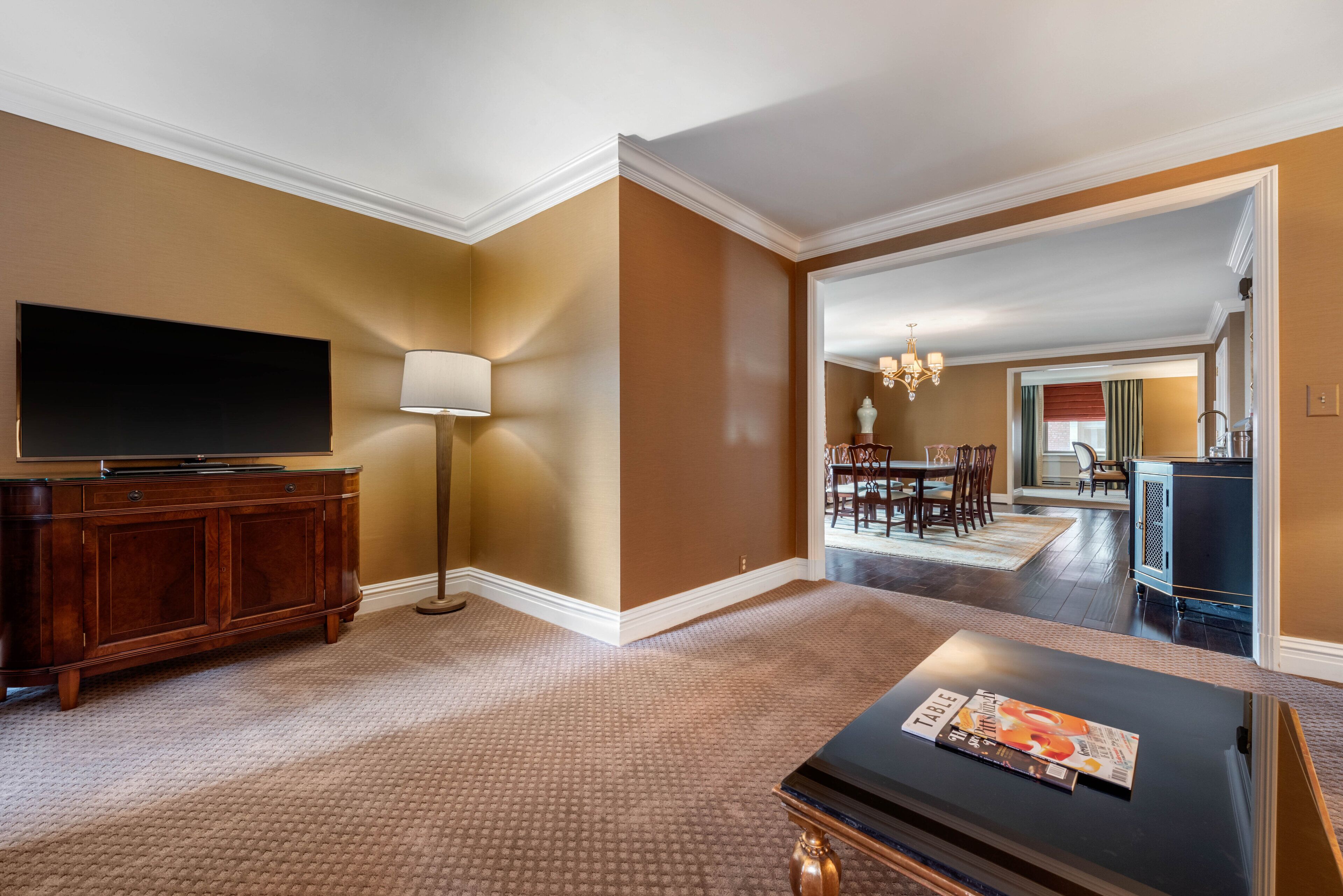 A hotel room with a flat-screen TV, a wooden cabinet, a lamp, and a dining area with a chandelier.