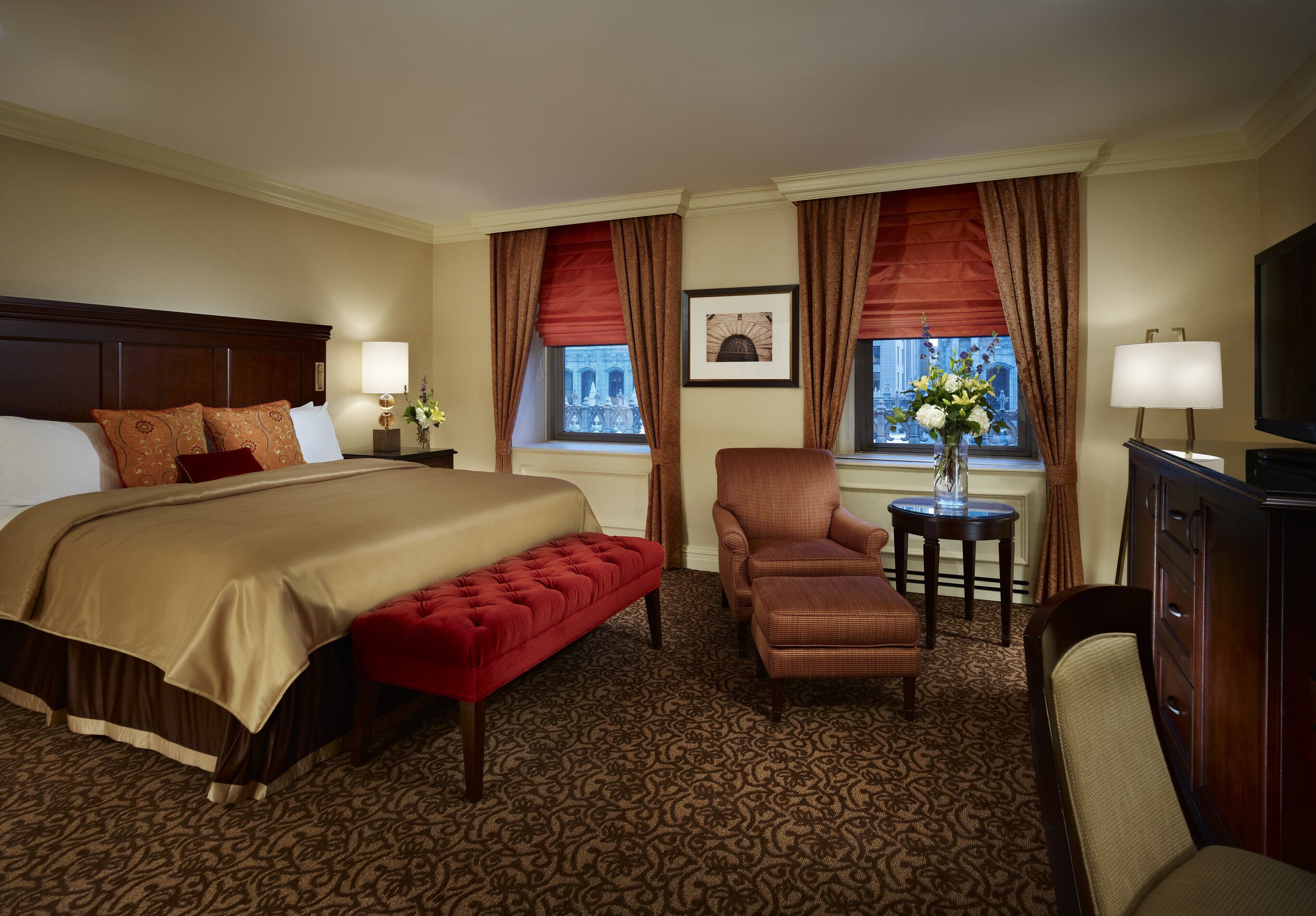A hotel room with a large bed, a red bench, a chair, a small table with a vase, and a television.