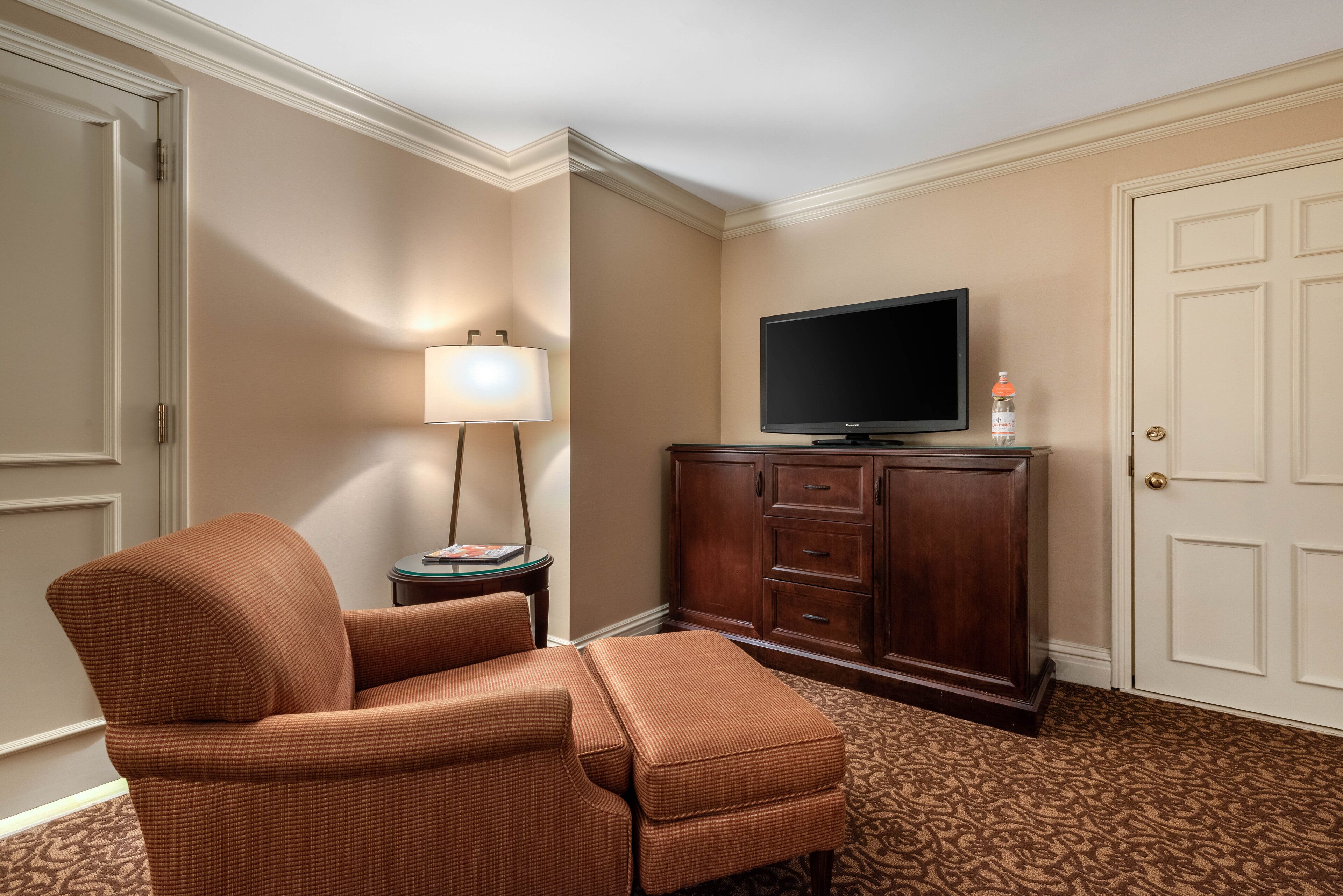 A hotel room with a brown upholstered armchair, a television on a wooden cabinet, a lamp, and a patterned carpet.