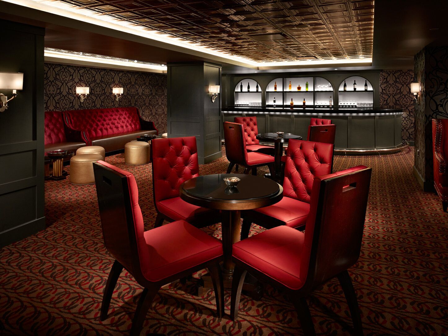 A dimly lit bar area with red upholstered seating, a patterned carpet, and a bar counter with bottles.