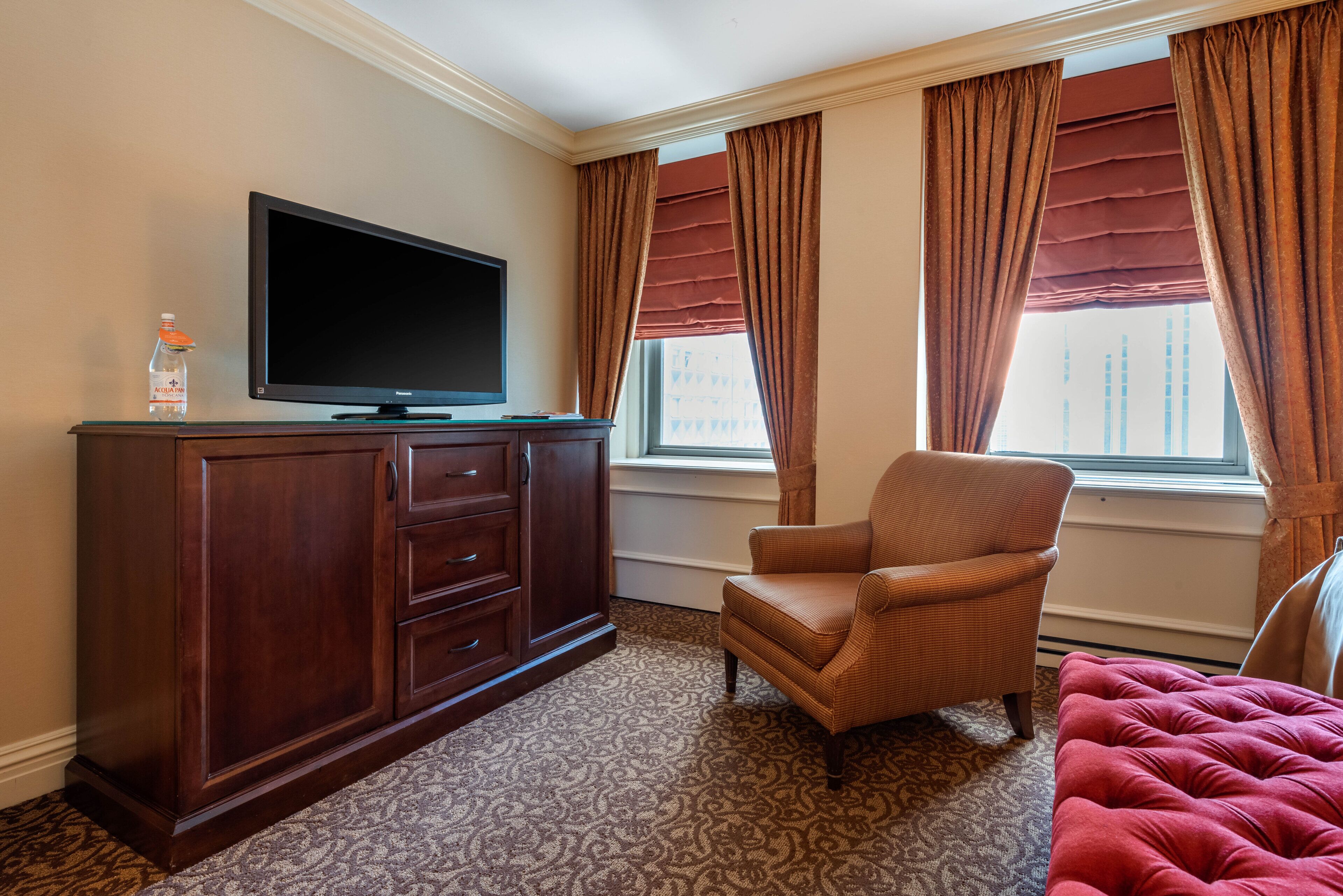 A hotel room with a flat-screen TV on a wooden cabinet, a patterned carpet, a red upholstered chair, and a large window with brown curtains.