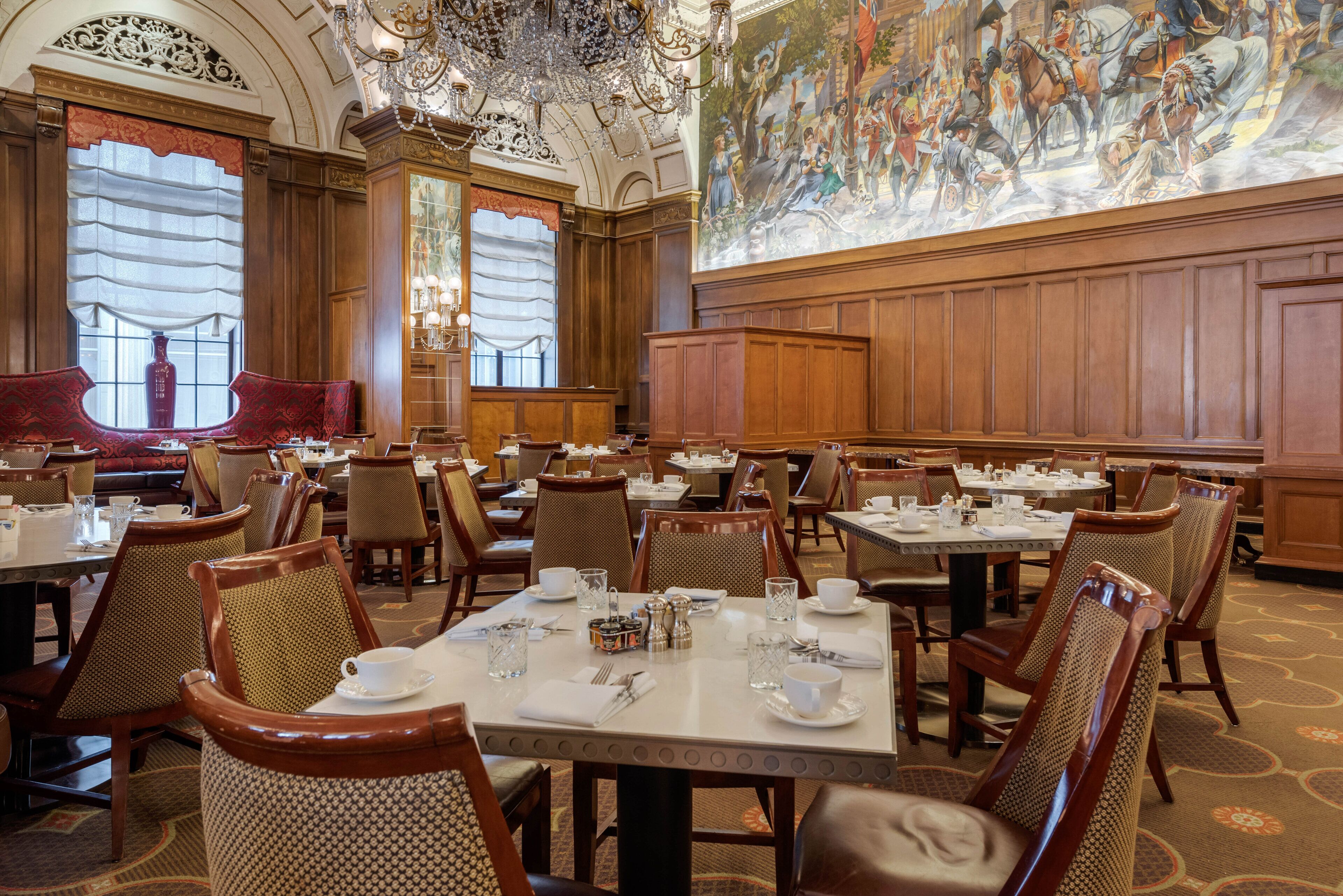 A formal dining room with multiple tables set for service, ornate chandeliers, and a large mural on the wall.