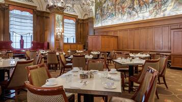 A formal dining room with multiple tables set for service, ornate chandeliers, and a large mural on the wall.