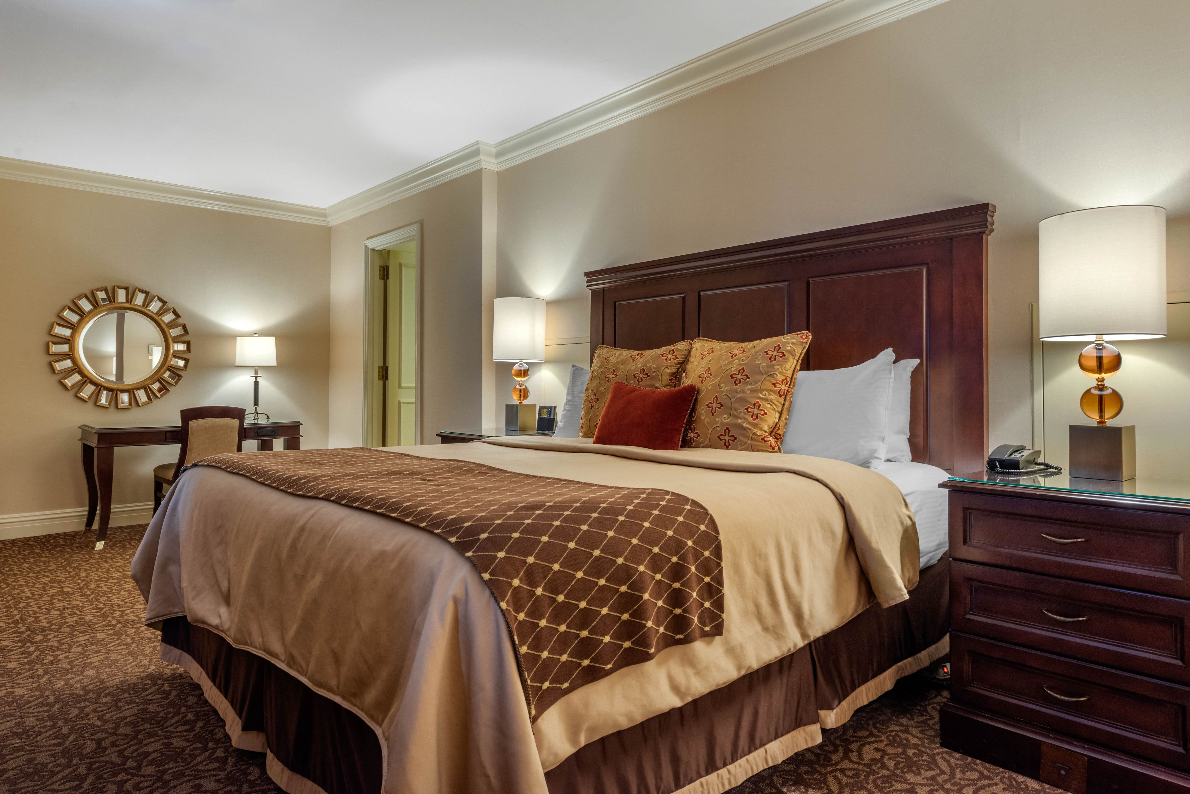 A hotel room with a large bed, a wooden headboard, a desk with a chair, two bedside lamps, and a mirror on the wall.