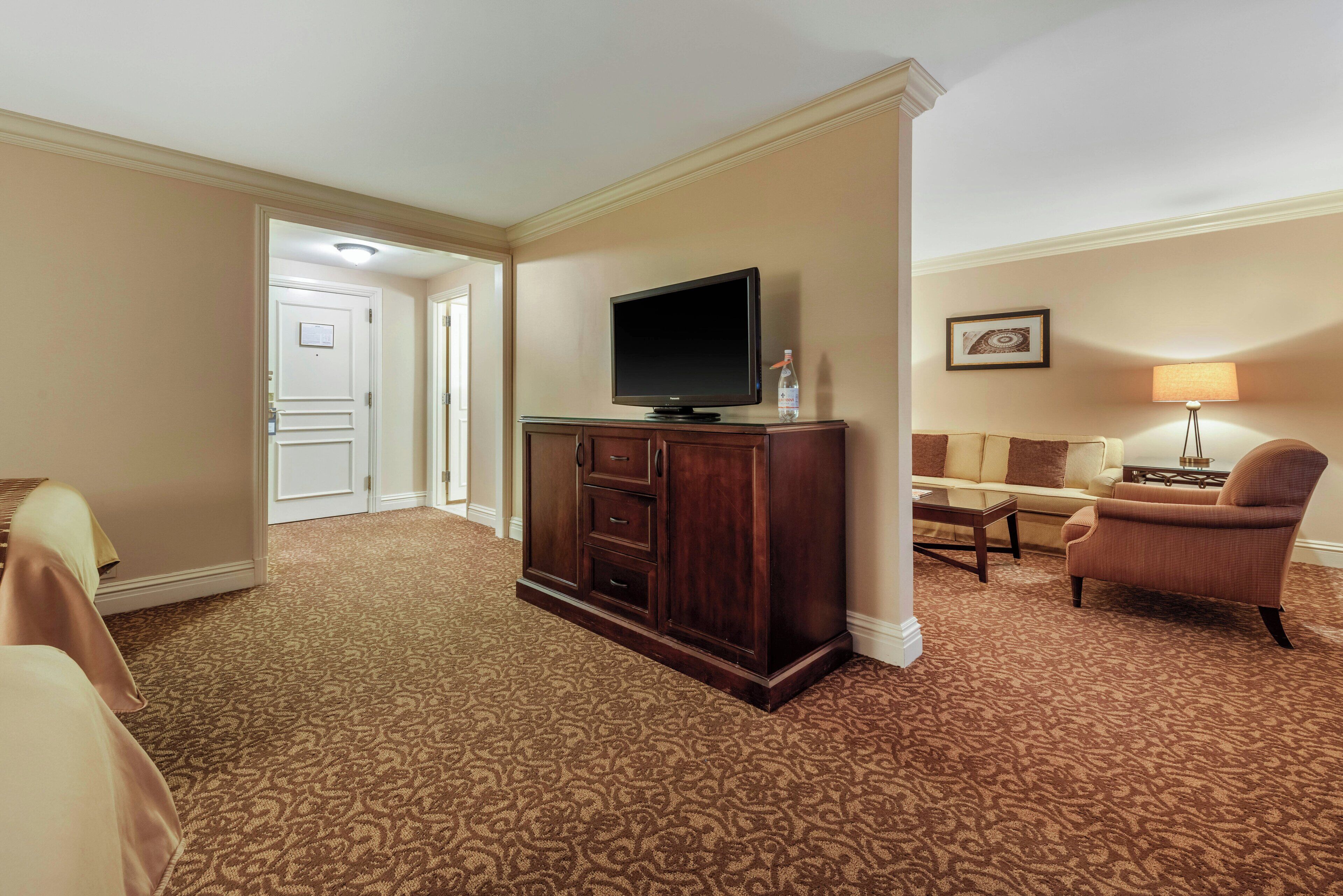 A hotel room with a large flat-screen TV on a wooden cabinet, a sofa, a chair, and a lamp.