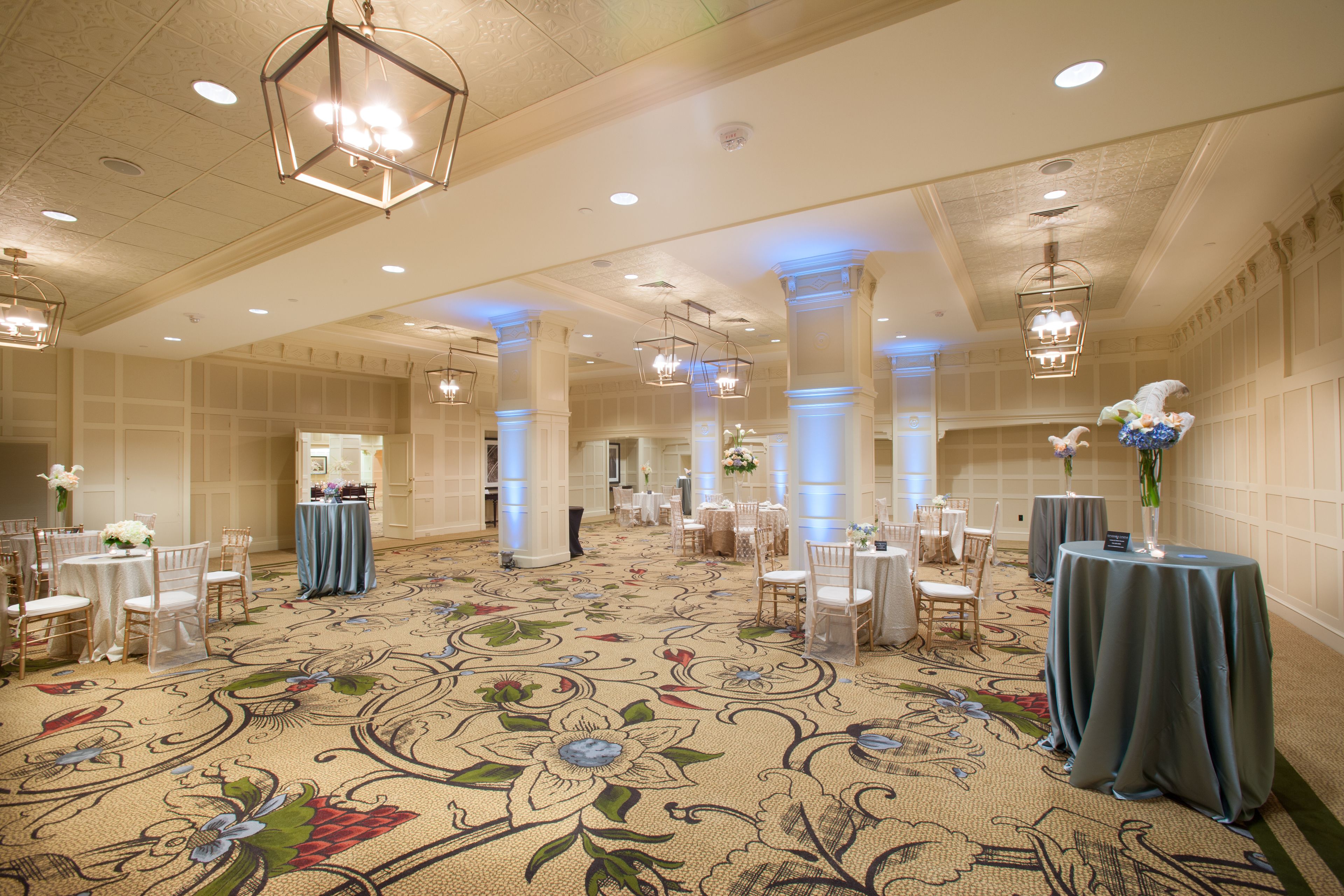 A spacious banquet hall with ornate floral carpet, multiple tables with blue tablecloths, and elegant chandeliers.