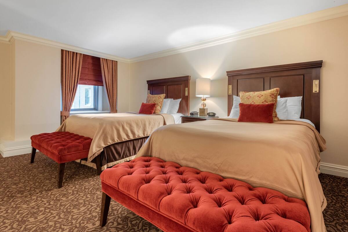 A hotel room with two beds, a red ottoman, a nightstand with a lamp, and a window with curtains.