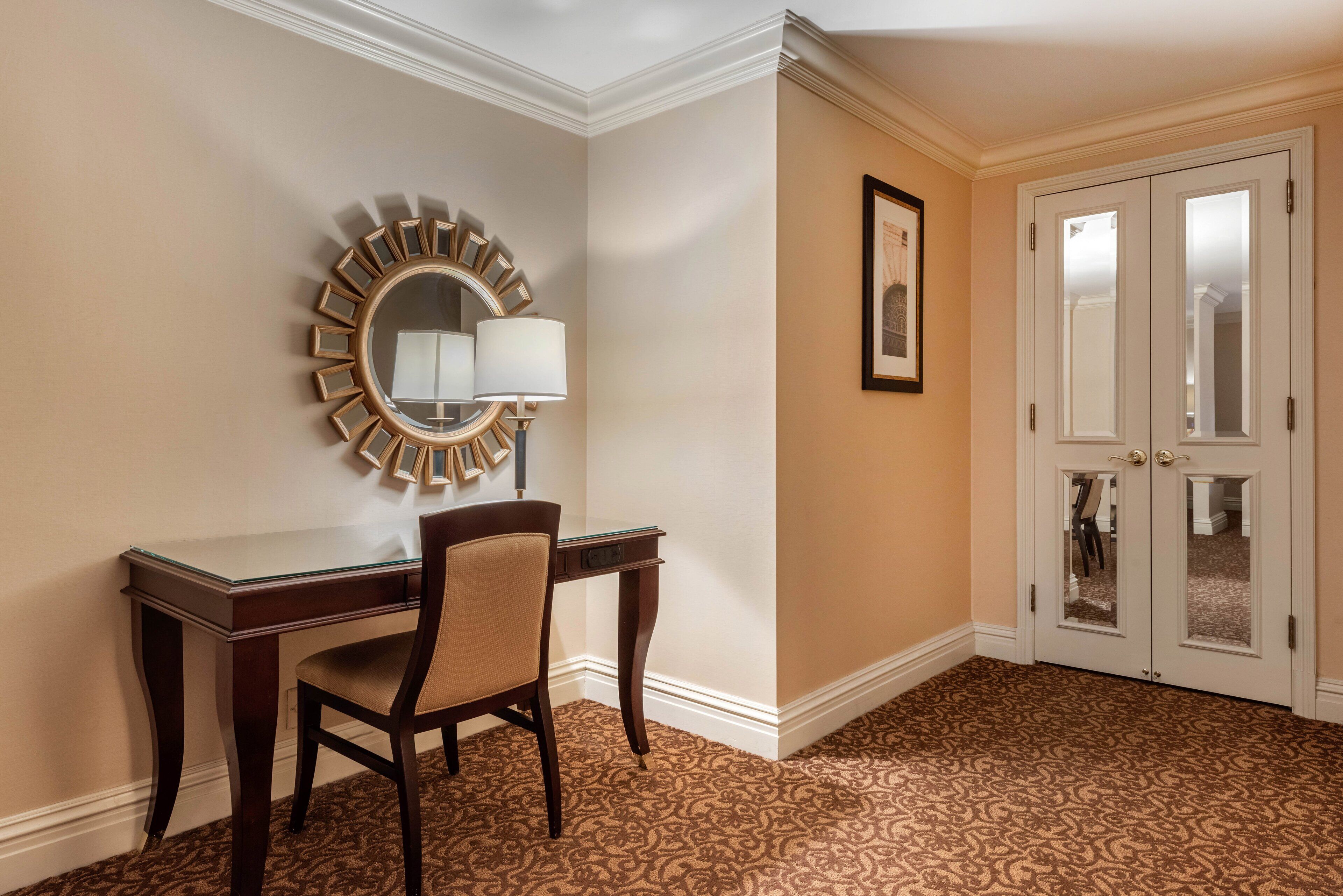 A hotel room with a desk, chair, a round mirror with a sunburst frame, a framed picture on the wall, and a pair of double doors.