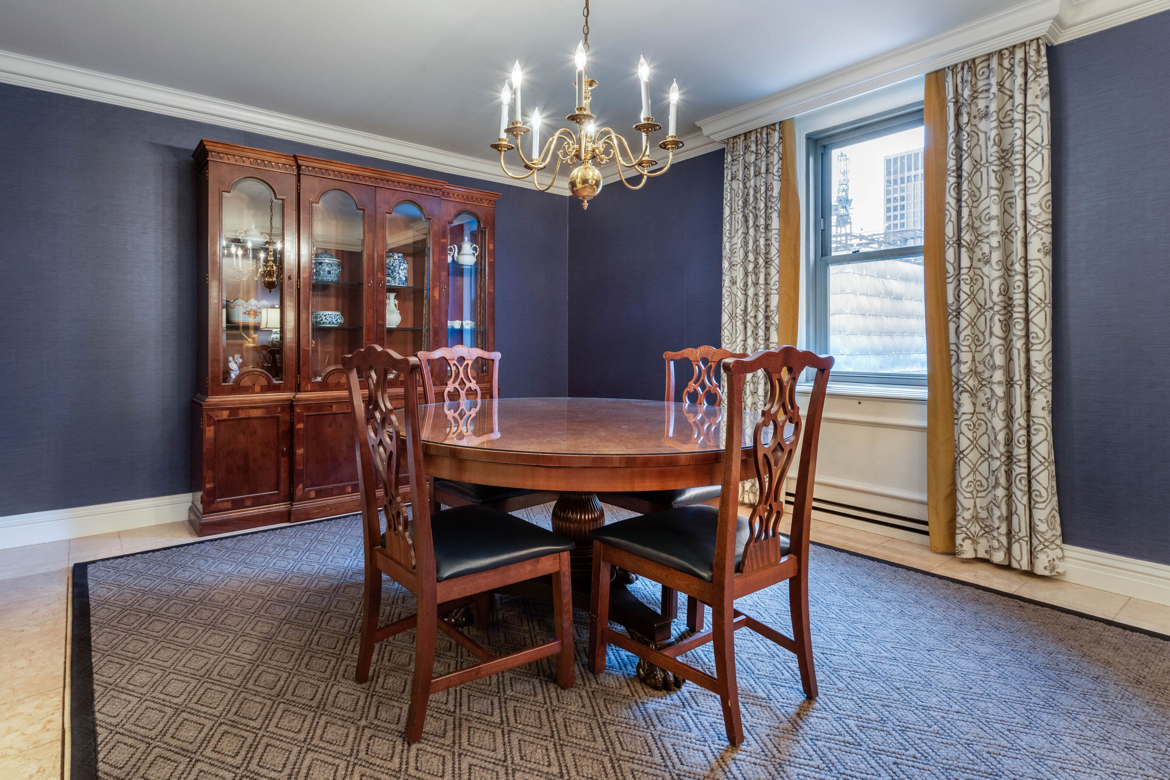 A dining room with a round table, four chairs, a chandelier, a wooden cabinet, and a window with curtains.
