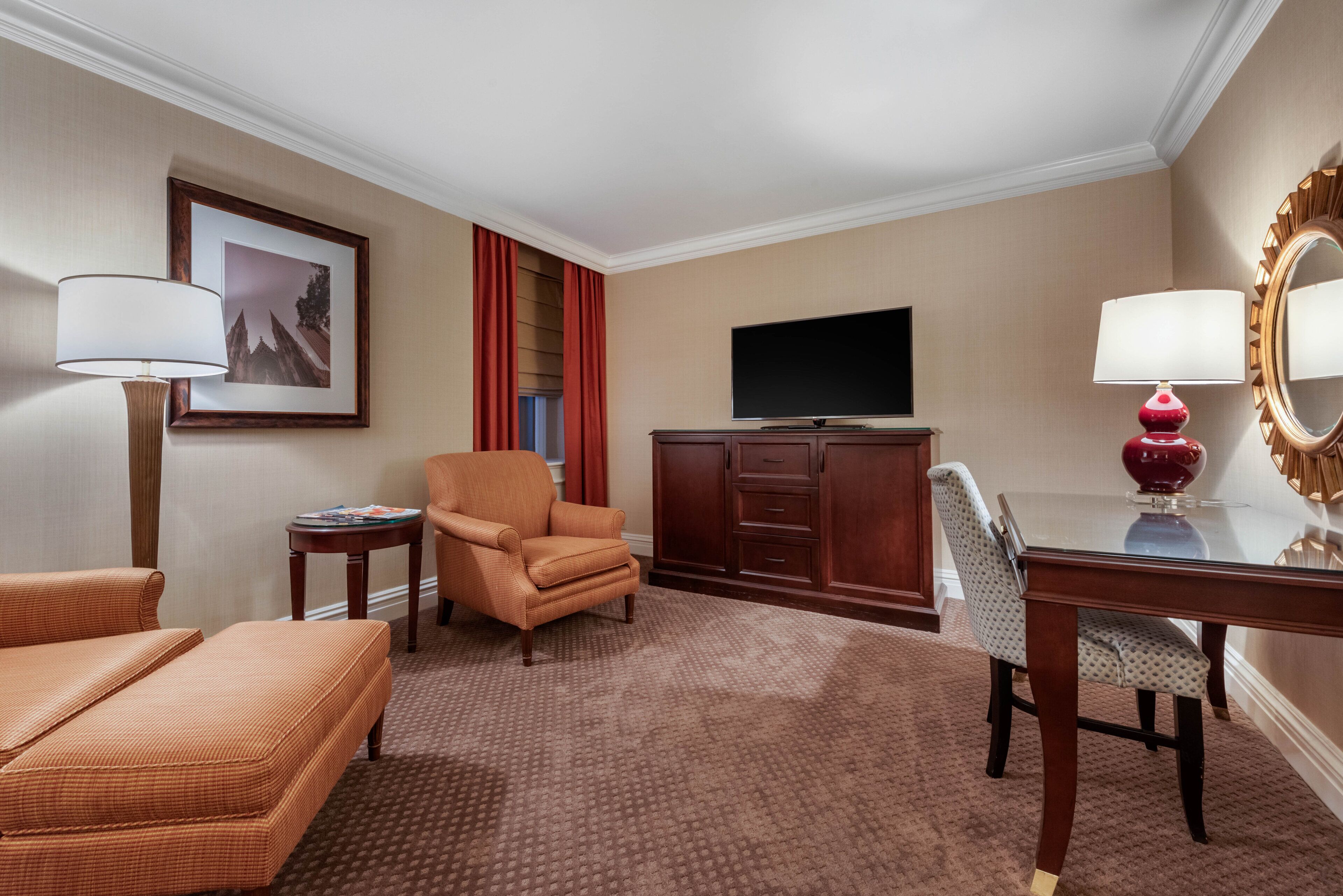 A hotel room with a sofa, armchair, TV, and a desk with a mirror.
