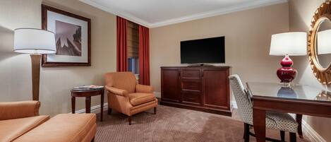 A hotel room with a sofa, armchair, TV, and a desk with a mirror.