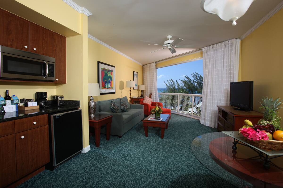 A compact hotel room with a kitchenette, a sofa, a dining table, and a balcony with a view.
