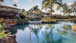 Zona de piscina en un resort con cascada, palmeras y un edificio con balcones.
