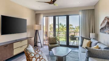 Una habitación de hotel moderna con un televisor de pantalla plana grande, un ventilador de techo, un balcón con vistas a palmeras y una zona de estar con sofá y sillones.