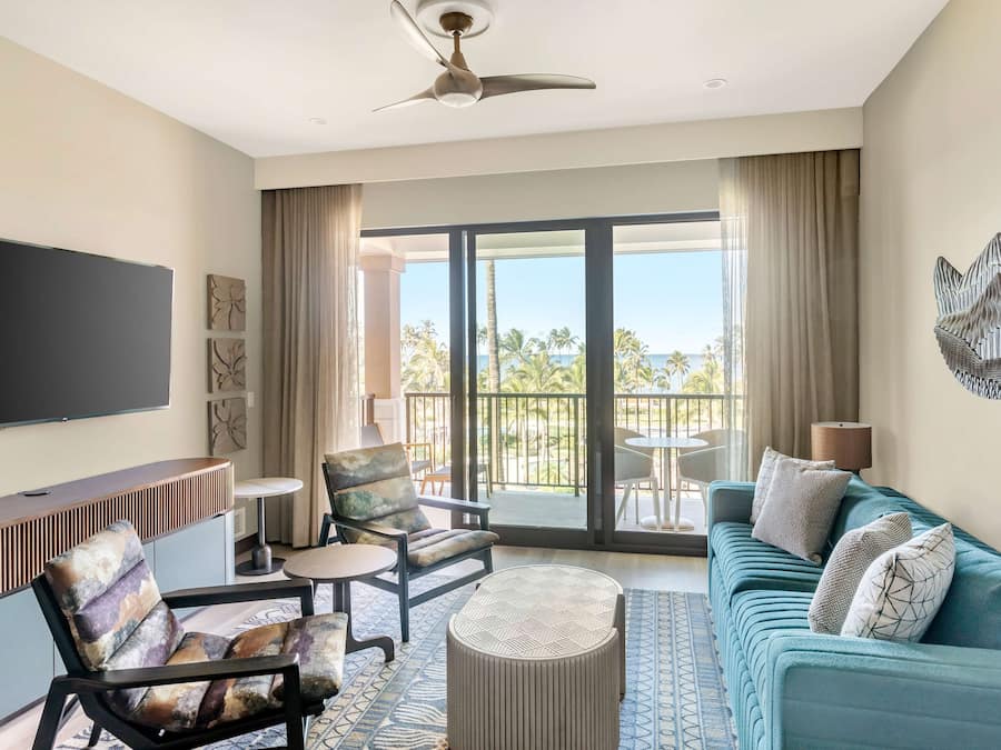 A hotel room with a large balcony, a sofa, a chair, a coffee table, and a TV.