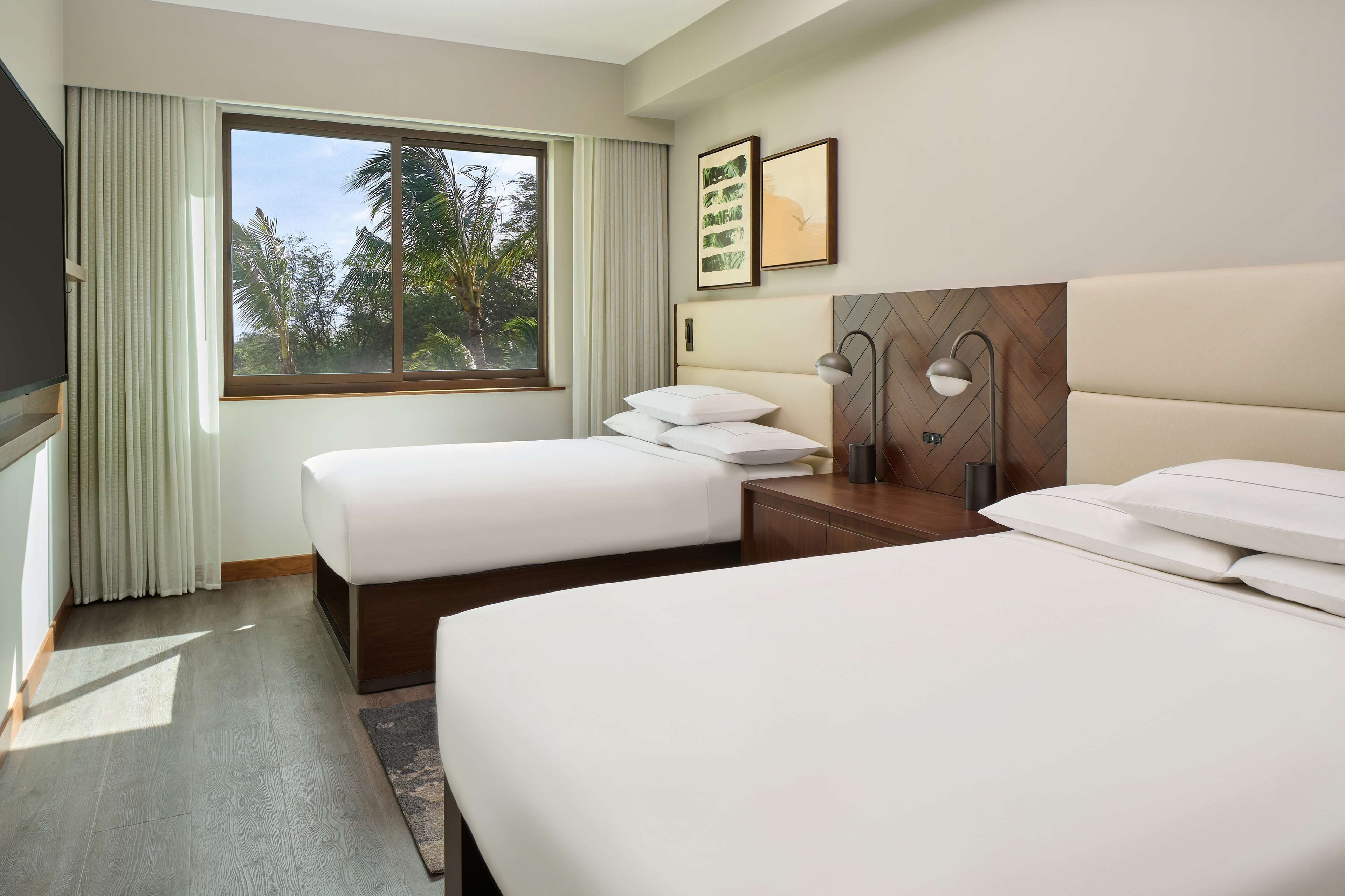 Une chambre d’hôtel avec deux lits, une grande fenêtre avec des rideaux, un téléviseur à écran plat et une tête de lit en bois.