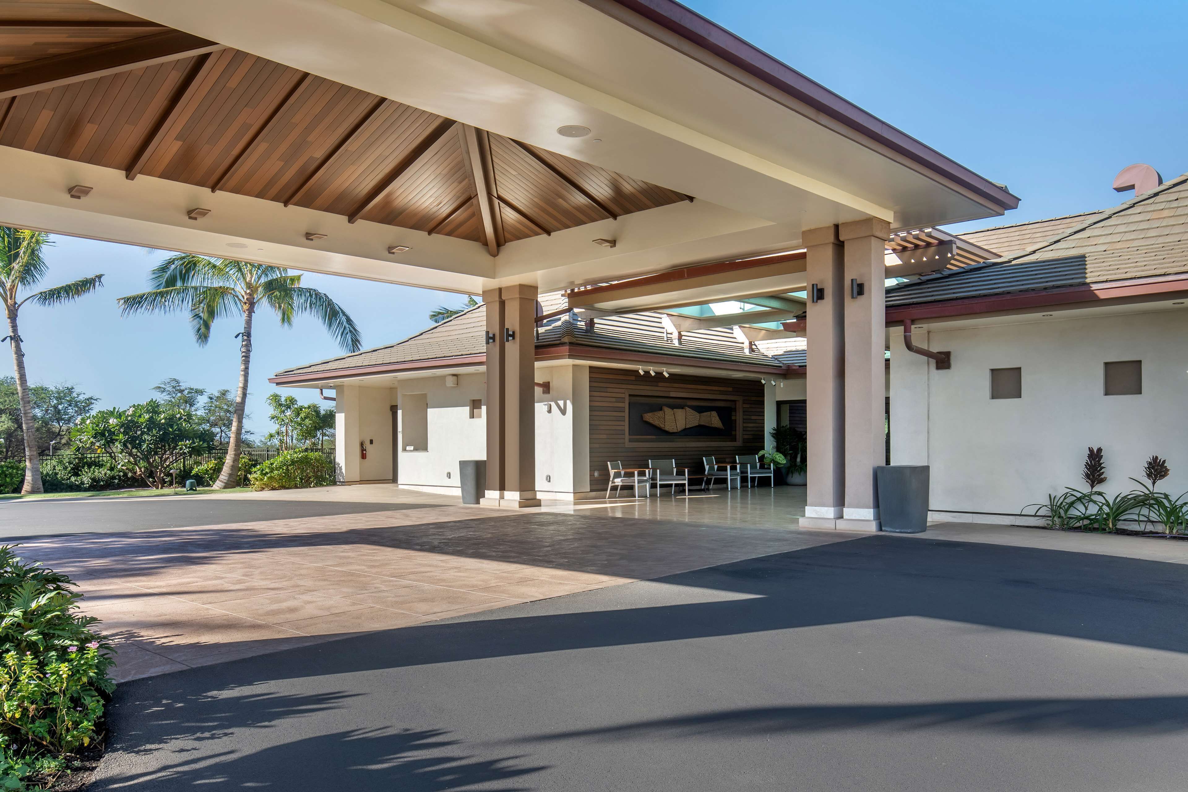 A covered parking area with a modern design, palm trees, and a building with a sloped roof.