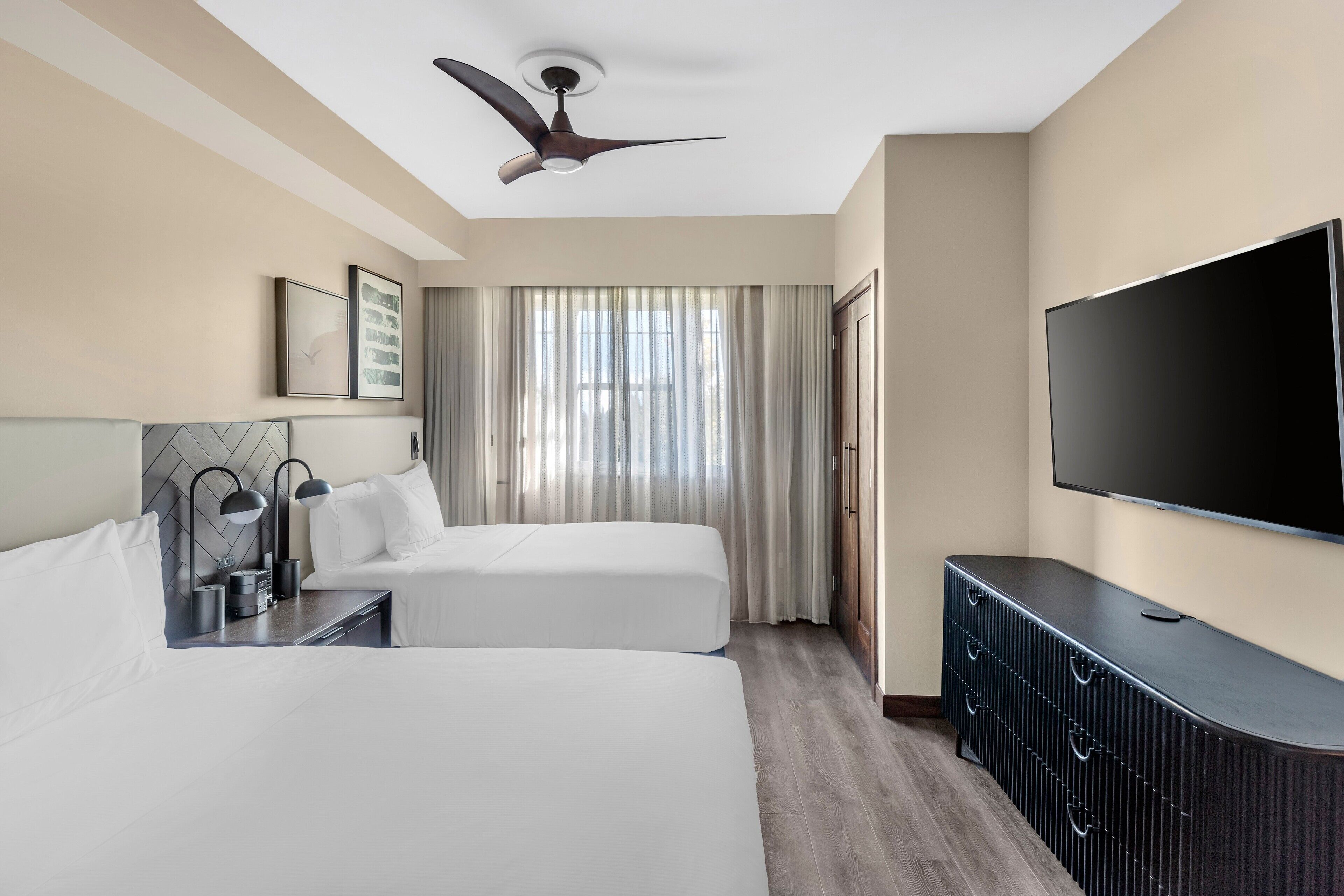 Quarto de hotel com duas camas, ventilador de teto, televisão e uma janela grande com cortinas.