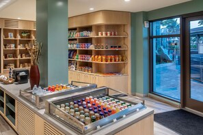 A hotel lobby with a self-service beverage station, shelves stocked with snacks and toiletries, and a view of the outdoor area through large windows.