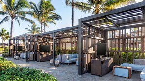 A covered outdoor seating area with sofas, tables, and planters, surrounded by palm trees.