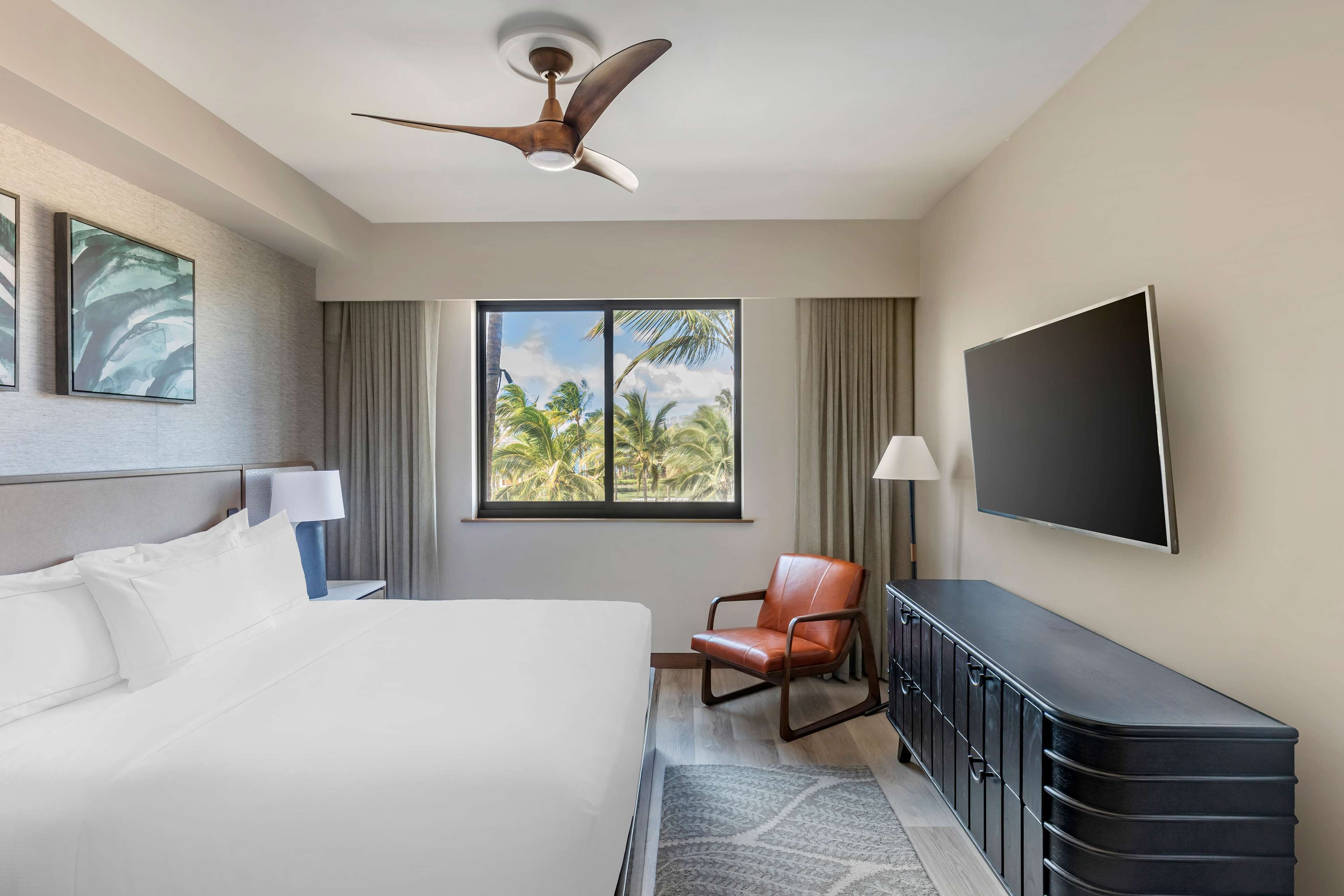 A hotel room with a bed, a desk, a chair, a television, and a window with a view of palm trees.