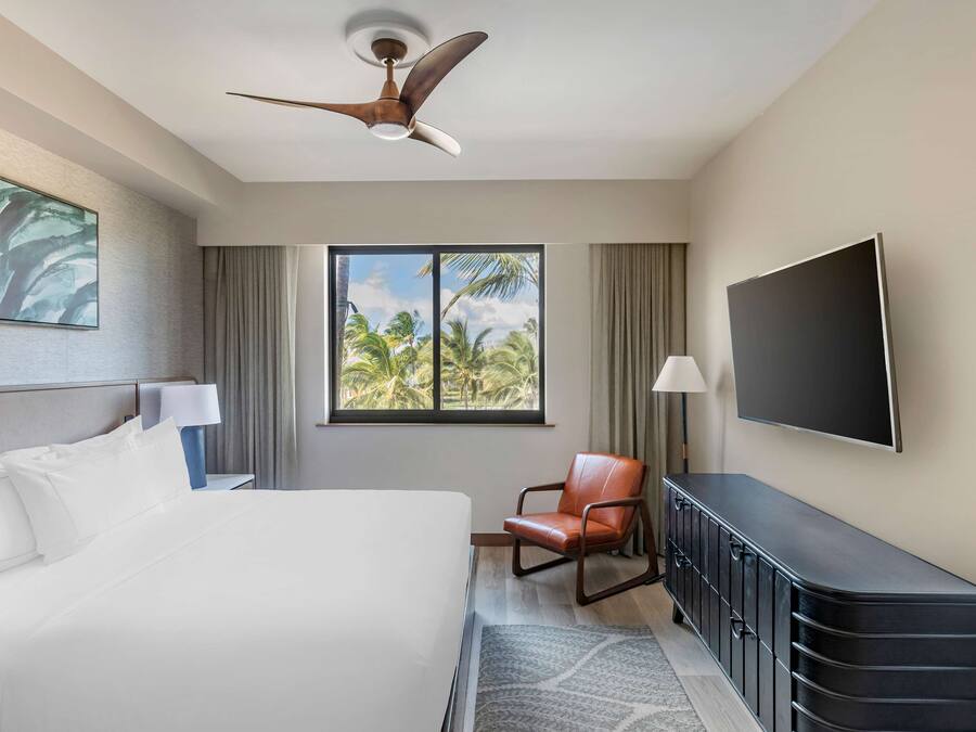 A hotel room with a bed, a desk, a chair, a television, and a window with a view of palm trees.
