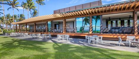 A modern building with a covered outdoor seating area, palm trees, and a view of the ocean.