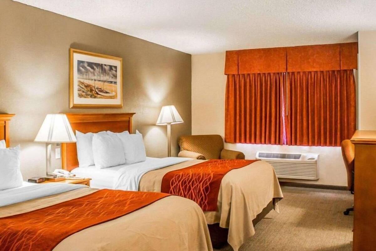 A hotel room with two beds, a desk, a chair, a lamp, an air conditioner, and a painting on the wall.