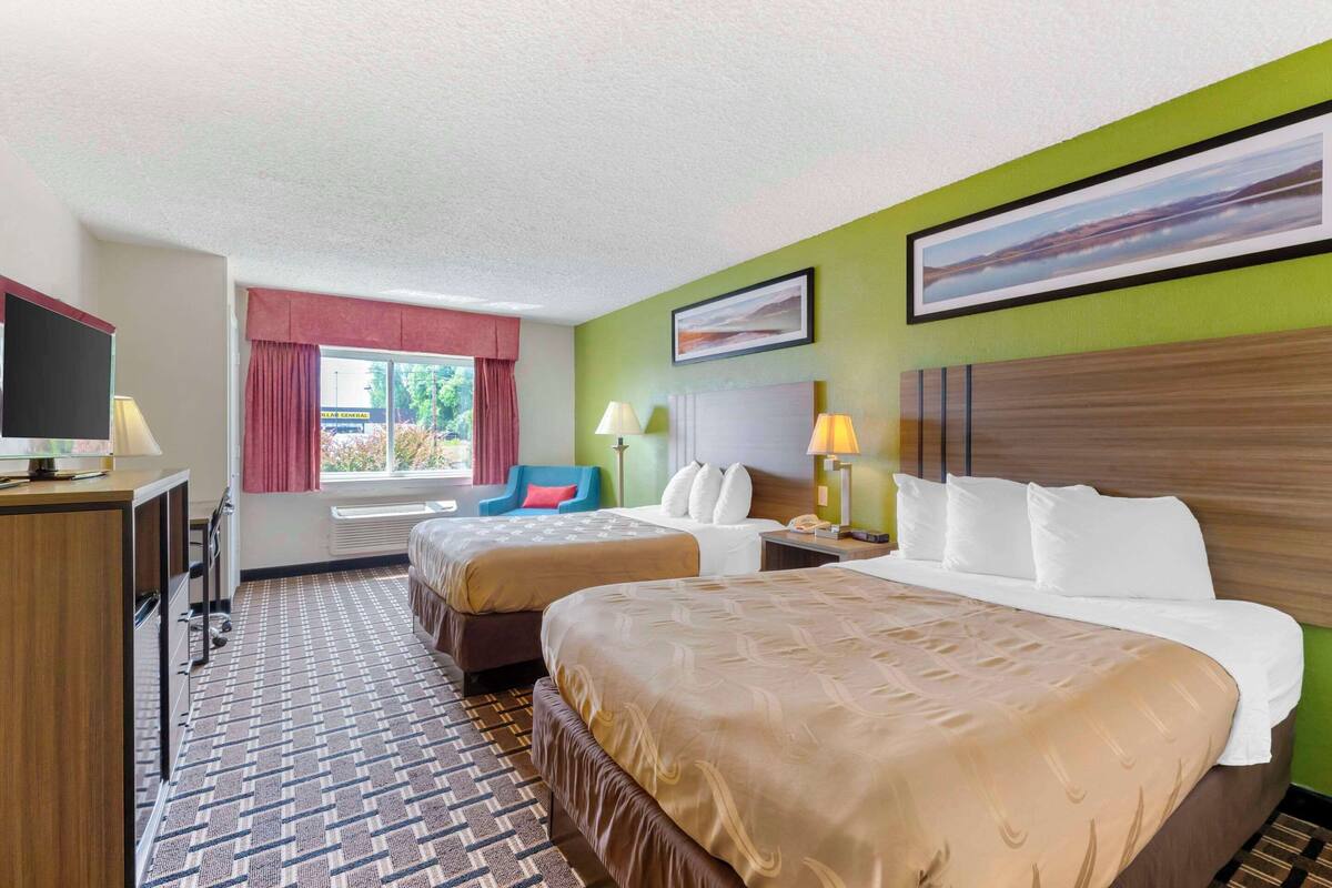 A hotel room with two beds, a desk, a TV, and a window with curtains.