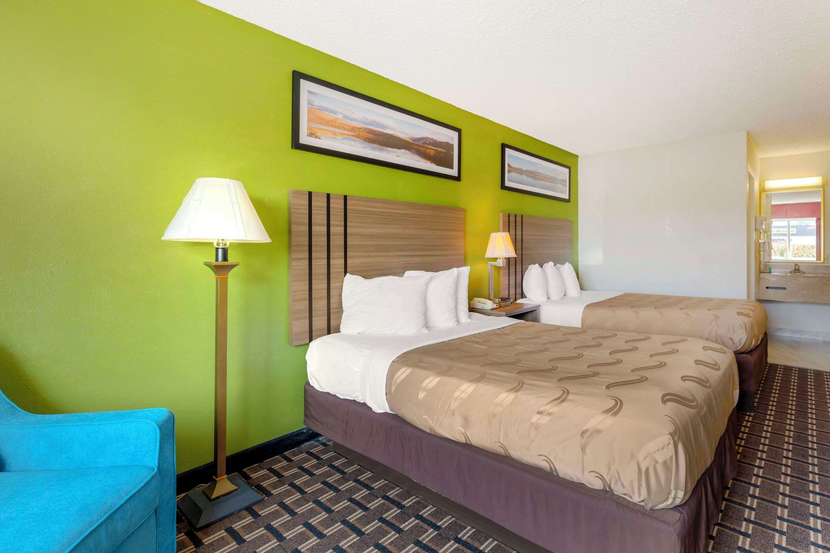 Ein Hotelzimmer mit zwei Betten, einem blauen Sessel, einer Stehlampe und zwei gerahmten Bildern an der Wand.