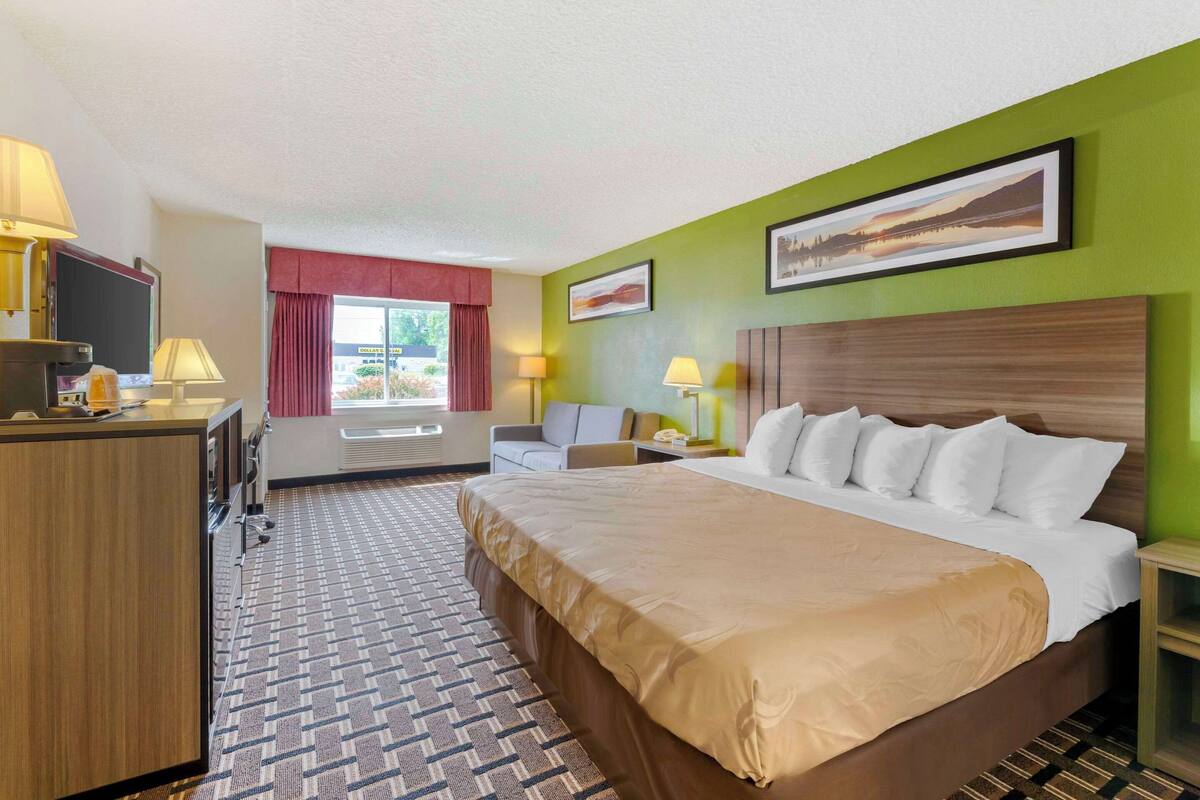 A hotel room with a large bed, a desk with a TV, a chair, and a window with red curtains.