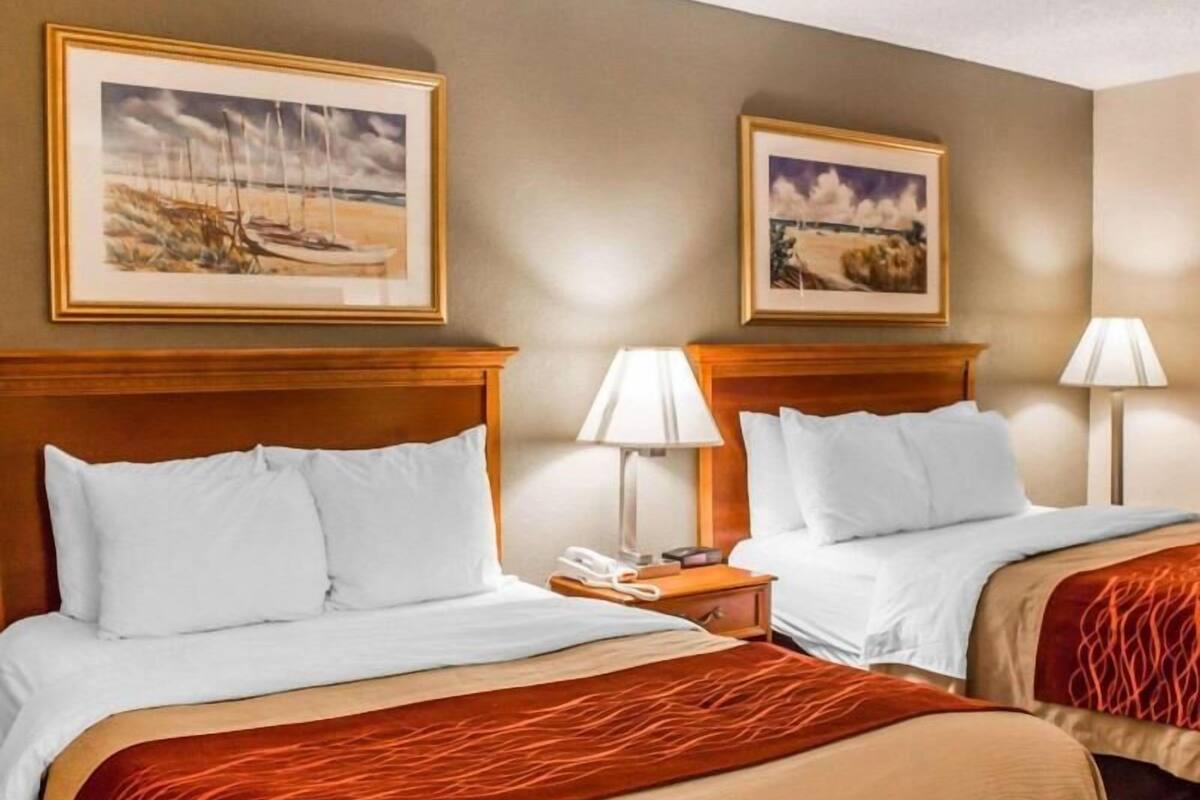 A hotel room with two beds, each with white bedding and a red accent. There are two framed pictures on the wall, a bedside table with a lamp, and a phone on the nightstand.