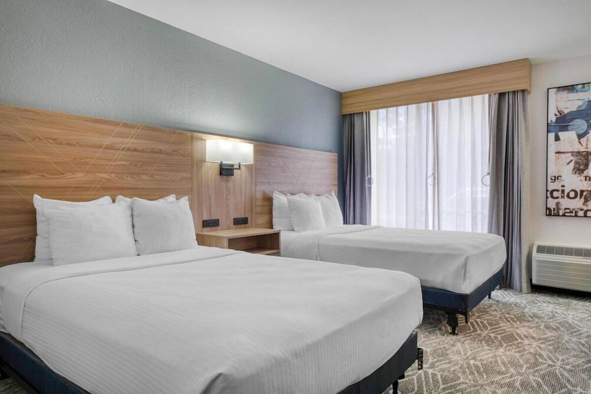 Habitación de hotel con dos camas, cabecero de madera, mesita de noche, lámpara de pared y un ventanal con cortinas.