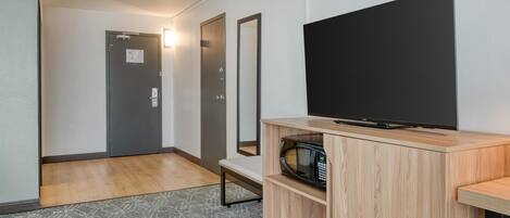 Habitación de hotel con una televisión de pantalla plana sobre un soporte de madera, un microondas, un banco y una puerta con una luz encima.
