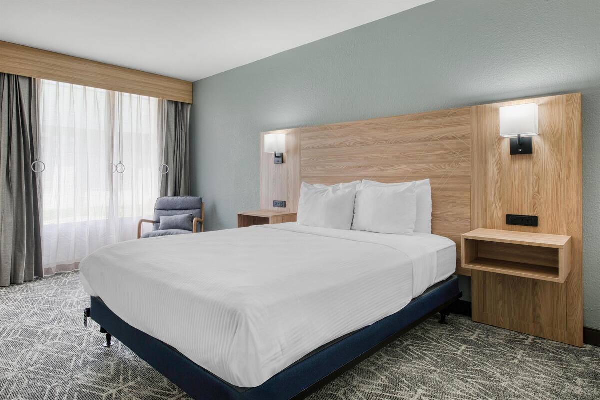 Una habitación de hotel con una cama grande, una silla, una mesita de noche y una lámpara fijada a la pared.
