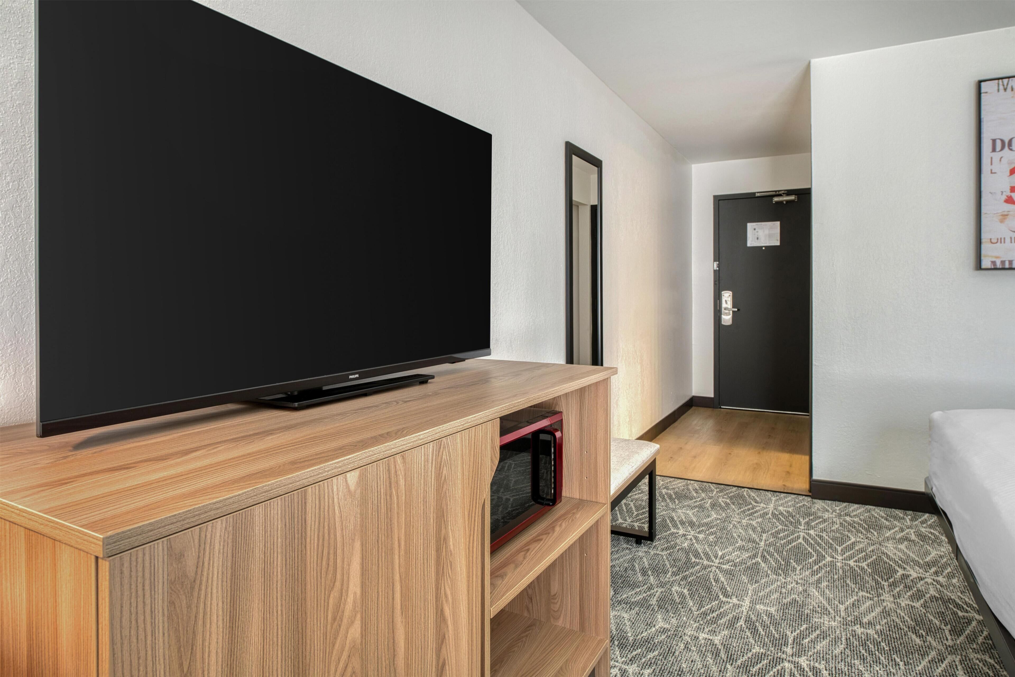 A modern hotel room with a large flat-screen TV on a wooden stand, a bed, a small seating area, and a door leading to another room.