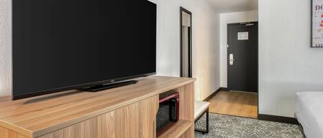 A modern hotel room with a large flat-screen TV on a wooden stand, a bed, a small seating area, and a door leading to another room.